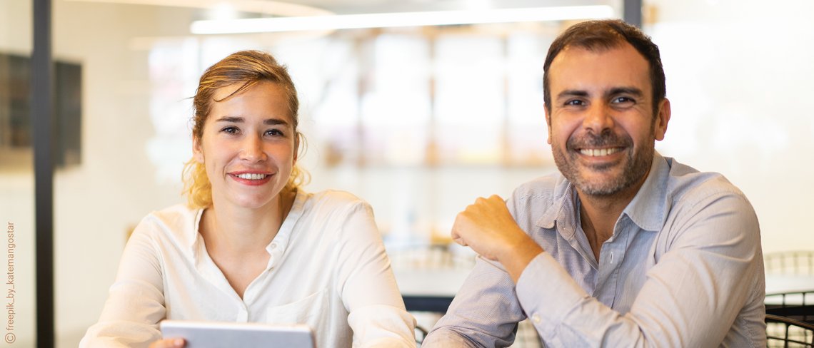 Zwei Personen sitzen an einem Tisch in einem hellen Büro mit Glaswänden und moderner Einrichtung.