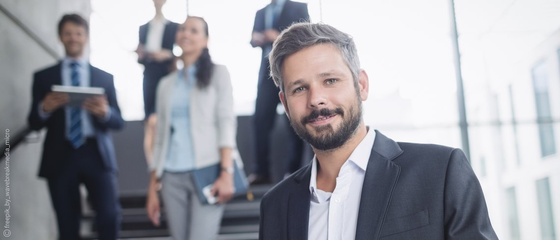 Eine Gruppe von Personen in formeller Kleidung, teils mit Tablets und Smartphones, auf einer Treppe in einem modernen Bürogebäude.