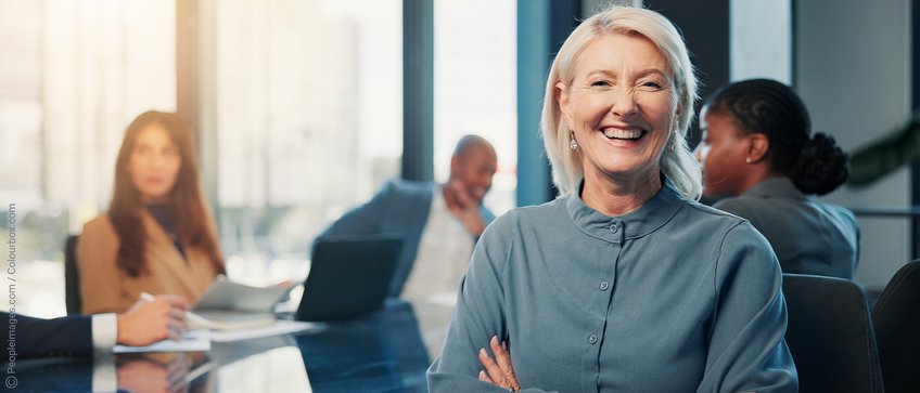 Gemeinsam Zukunft gestalten – werden Sie Teil unseres Netzwerks In einem hellen Besprechungsraum sitzen Mitarbeiter an einem Tisch, konzentriert auf ihre Aufgaben, während eine Frau im Vordergrund in die Kamera blickt.