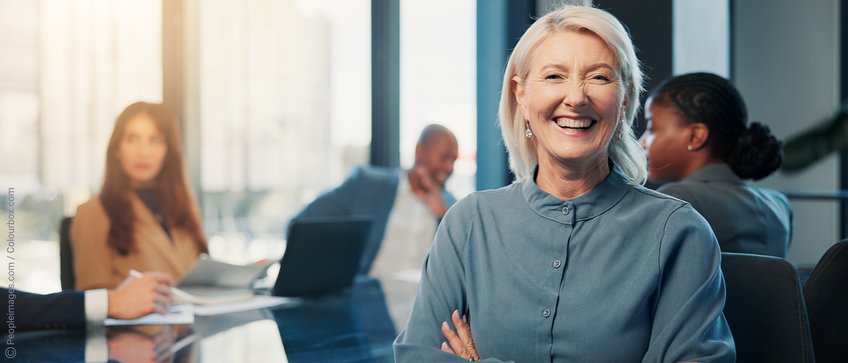 Shaping the Future Together — Join Our Network In a brightly lit conference room, employees are seated around a table, focused on their work, while a woman in the foreground looks into the camera.